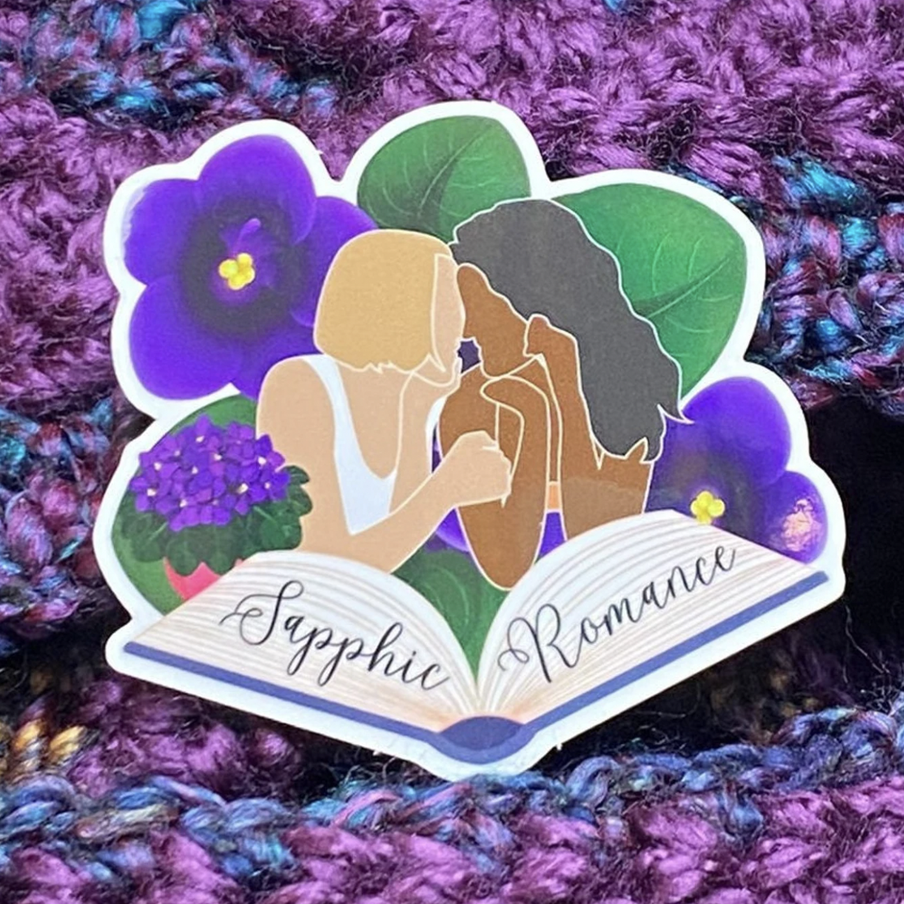 Image of a sticker with illustrations of two femmes kissing with pansies in the background and an open book at the bottom that says "sapphic romance"