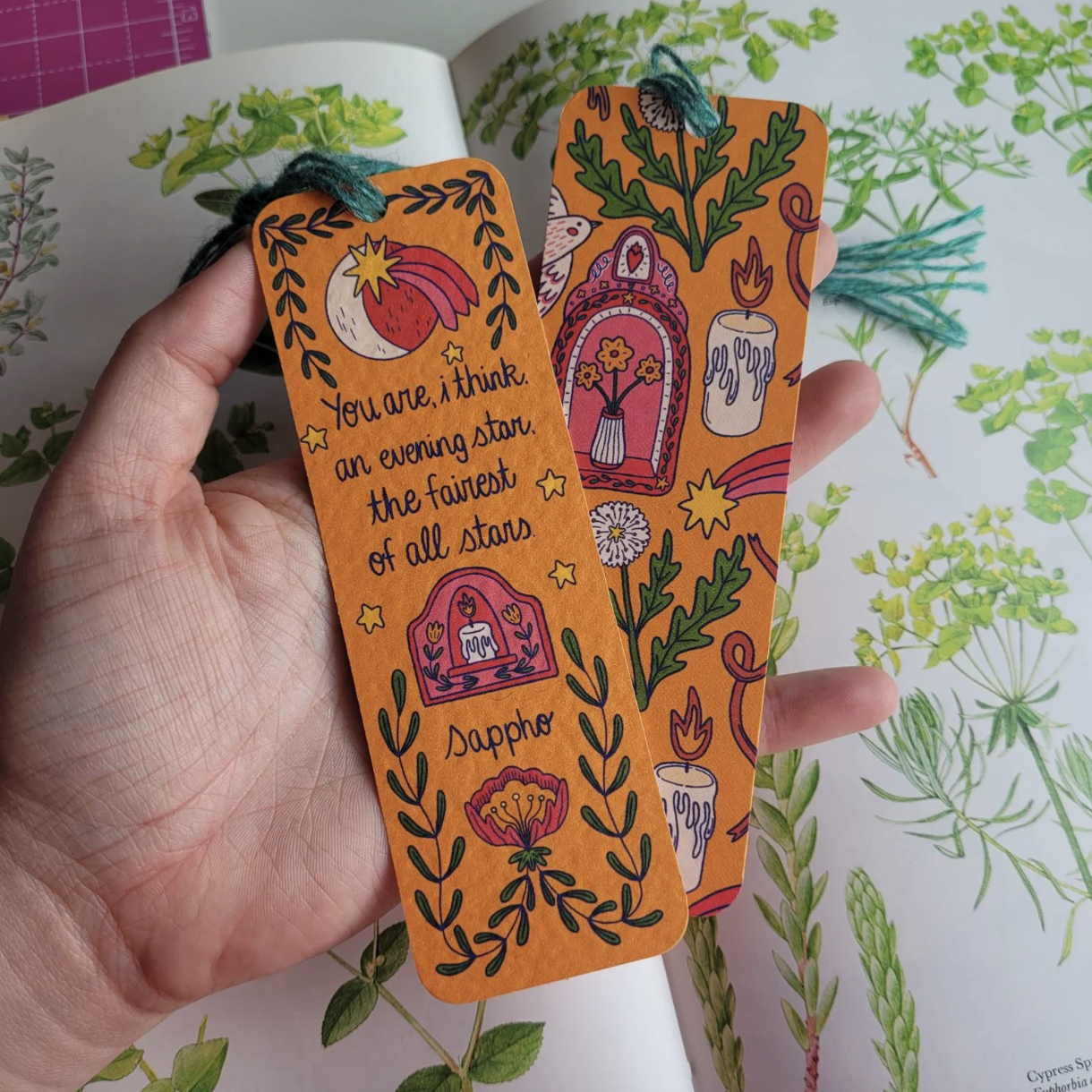 Image of a hand holding two bookmarks with Grecian inspired illustrations and a line from a Sappho poem: "You are, I think, an evening star, the fairest of all stars"