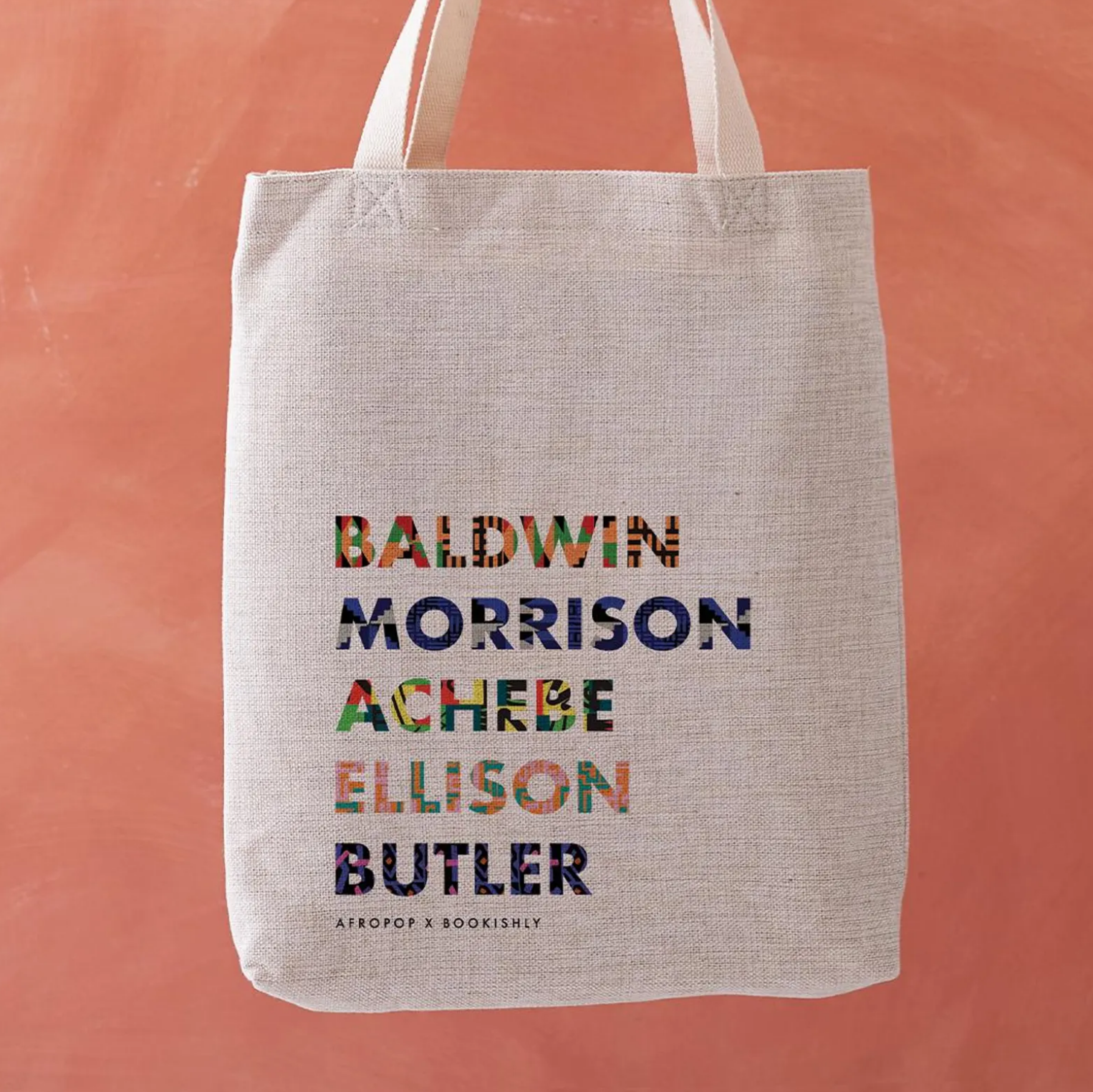 A white tote bag set against an orange background with colorfully screen-printed names of famous Black authors "Baldwin, Morrison, Achebe, Ellison, and Bulter"