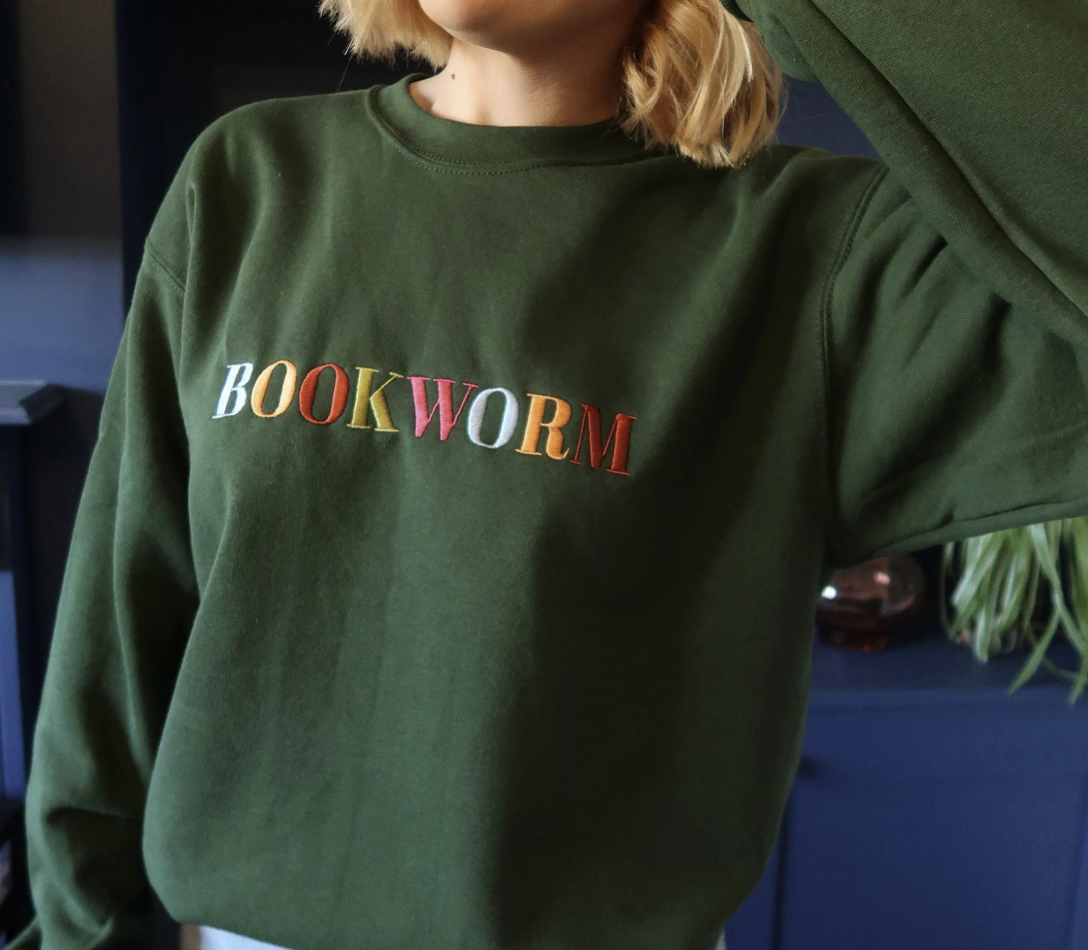 Picture of a white woman wearing a dark green sweatshirt embroidered with the word "bookworm" in a rainbow of colors across the chest