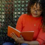 a tanned skin woman of color reading a book in a woven chair