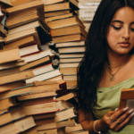 fab tan-skinned woman with long, dark hair and long nails is reading a book while sitting near huge stacks of books