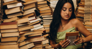 fab tan-skinned woman with long, dark hair and long nails is reading a book while sitting near huge stacks of books