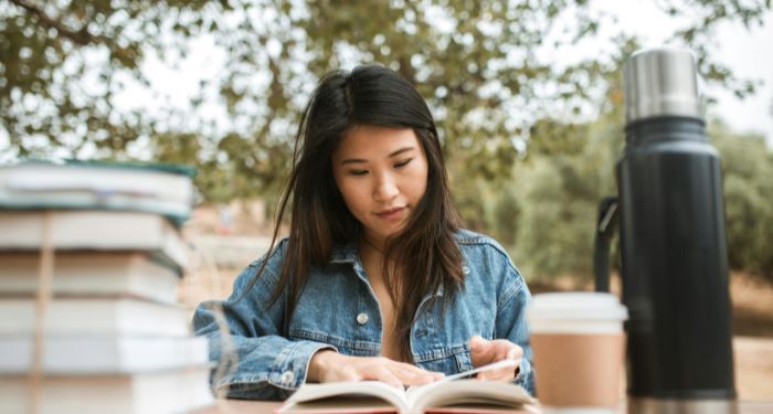 light-skinned Asian woman reading at a table with a thermos and a coffee cup