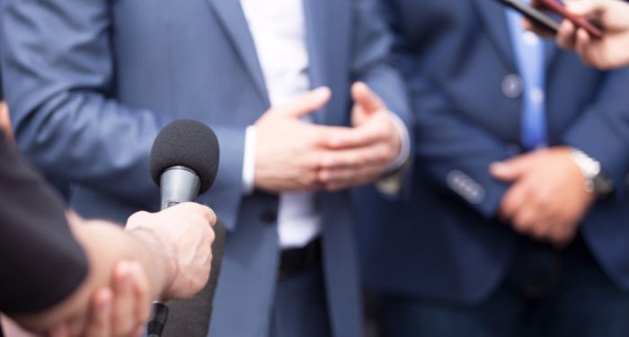 people in suits seen from the chest down. one person is gesturing with their hands, the others are holding microphones up to them