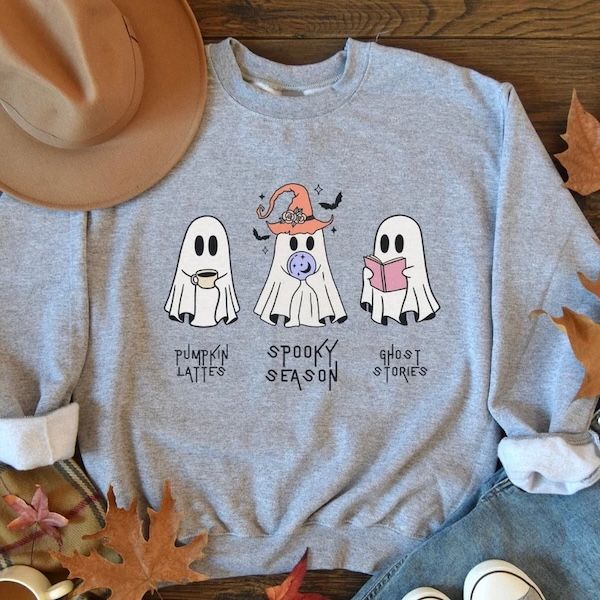 heathered grey crewneck sweatshirt with a graphic of three ghosts on the front. One is holding a coffee mug, one is wearing a pointy orange hat and holding a crystal ball, and one is holding a book. Beneath each ghost is corresponding text: pumpkin lattes, spooky season, and ghost stories