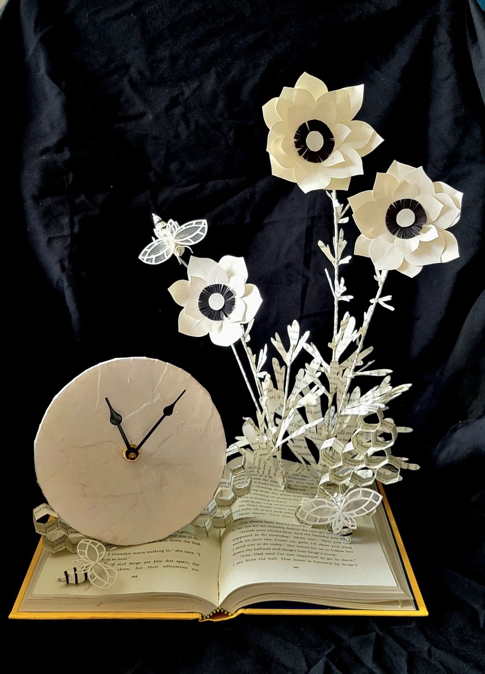 A book sculpture with flowers and a clock coming out of a book in front of a black background.