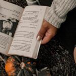 a brown-skinned hand holding open a book on the ground; there is a pumpkin under the book