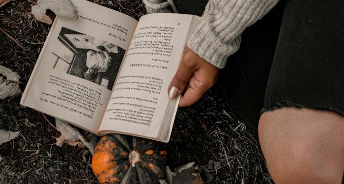 a brown-skinned hand holding open a book on the ground; there is a pumpkin under the book