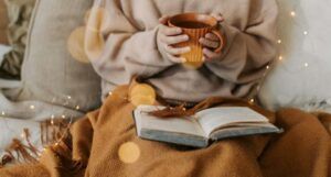 a fair-skinned person holding a mug with a book and autumn-themed decor in their lap