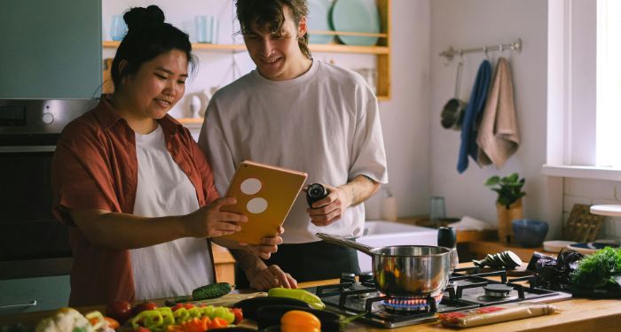 an Asian woman and white man looking at a tablet and cooking in a kitchen