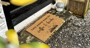 a doormat outside a door on gravel. doormat reads "this house runs on coffee and books"