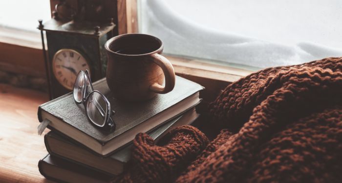 a coffee mug resting on a pile of books next to a blanket by a window with snow outside