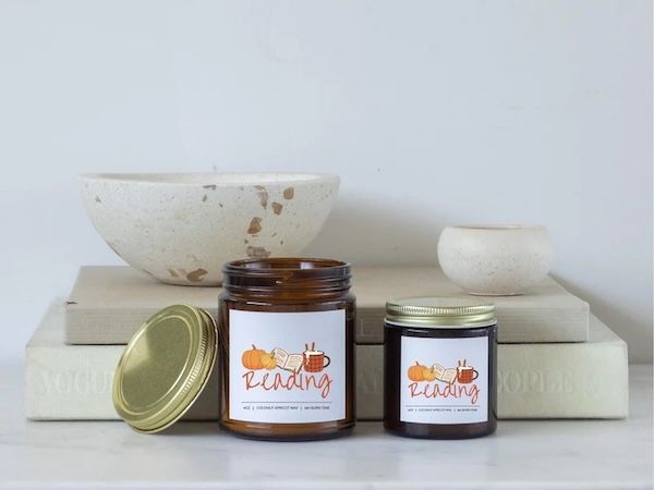 two amber glass candles with brass lids arranged in front of a small stack of books. The candles have grey labels with graphics of candles, books, and a coffee mug and text that says "Reading"