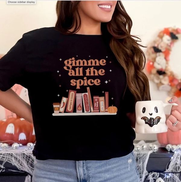 woman wearing a black tshirt with a graphic of several book spines bookended by a paper coffee cup and a pumpkin. Text hovering over the image reads "gimma all the spice"