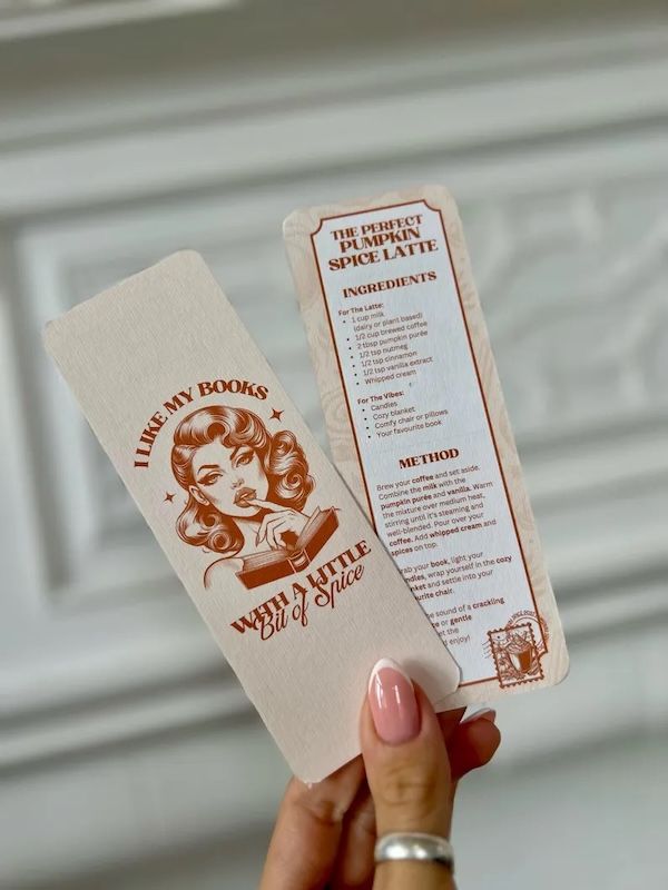 cream bookmark with text and design in reddish brown ink. The graphic is of a glamorous woman touching her lips with one finger with an open book in front of her. Text bordering the image reads "I Like My Books With a Little Bit of Spice"