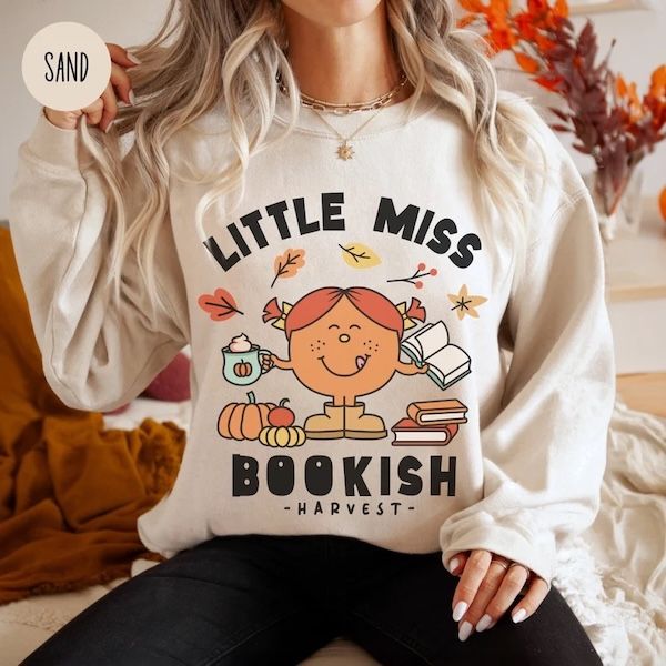 woman wearing a sand-colored crewneck sweatshirt with text that reads "Little Miss Bookish Harvest" bordering a graphic of a smiling girl holding a mug and a book