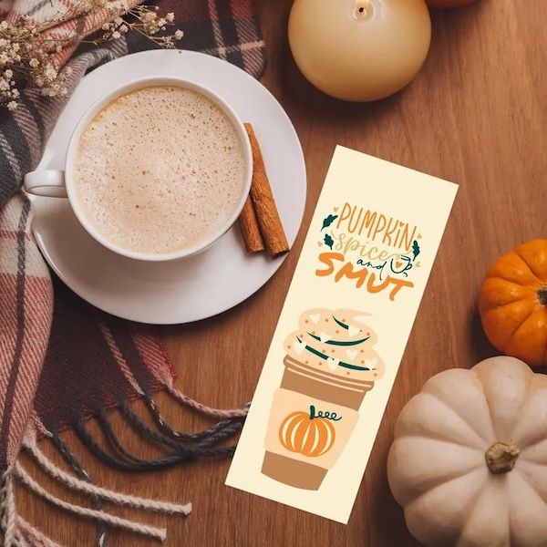 a cream bookmark resting next to a cup of frothy coffee. The bookmark contains a graphic of a coffee drink in a paper cup with a pumpkin on the sleeve and text that reads "Pumpkin Spice and Smut"