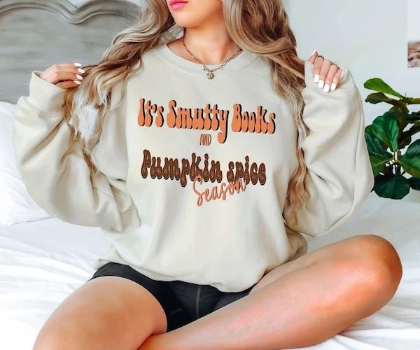 woman wearing a sand-colored crewneck sweatshirt with orange and brown text on the front that reads "It's Smutty Books and Pumpkin Spice Season"