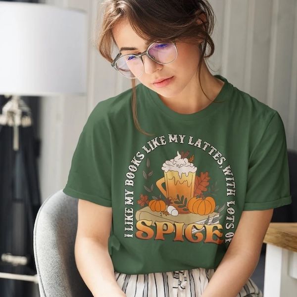 woman wearing a green tshirt with a design of a mug overflowing with whipped cream resting on top of a book between pumpkins. Text bordering the graphic reads "I LIie My Books Like My Lattes, with Lots of Spice"