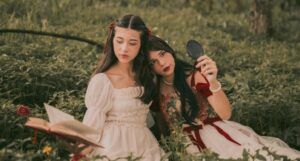 two fair-skinned woman sitting in a field next to each other; one is reading