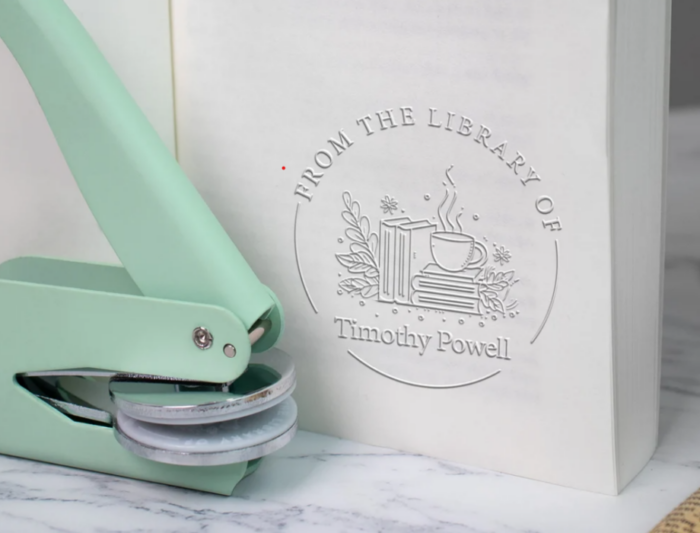 A metallic embosser beside a chunk of paper embossed with a "from the library of' message