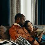 a dark brown-skinned Black man and a fair-skinned white woman reading a book on a couch