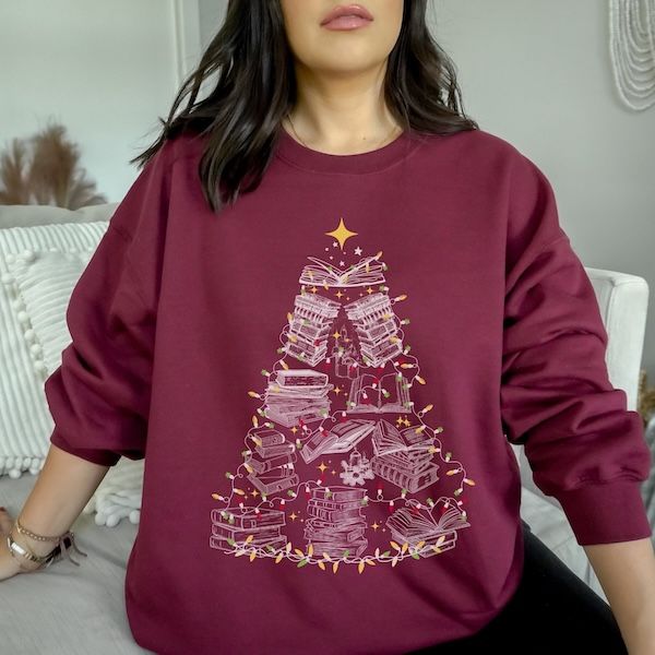 a white woman wearing a maroon crewneck sweater with a white stenciled design os books and lights arranged to form a Christmas tree shape