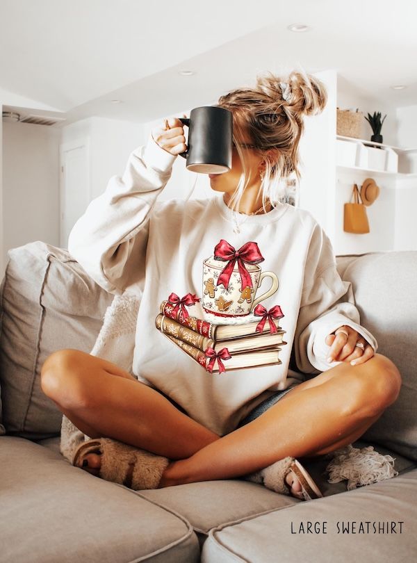 a white woman wearing a cream crewneck sweater with a graphic of a mug resting on a stack of three books. The mug has small gingerbread men and snowflakes on it, and the books and mug are also adorned with red bows