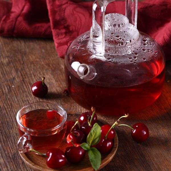 amber-colored tea in a clear tea pot next to a cup of tea on a saucer containing a handful of cherries