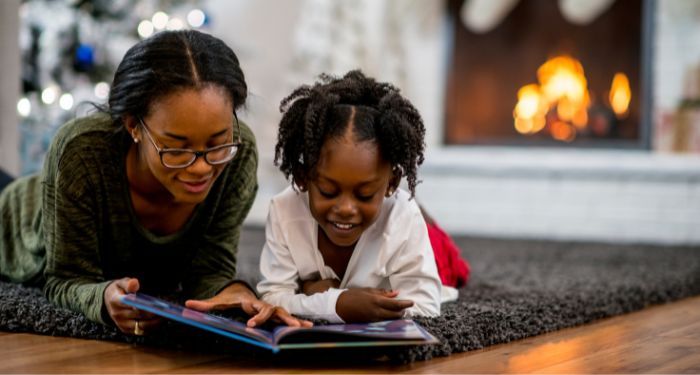 A Black woman and child reading a picture book on the floor of a living room with a Christmas tree and fireplace in the background