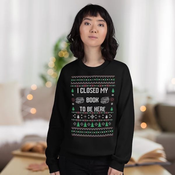 an Asian woman in a black crewneck sweater in the ugly Christmas sweater style with text that reads "I Closed My Book to Be Here"