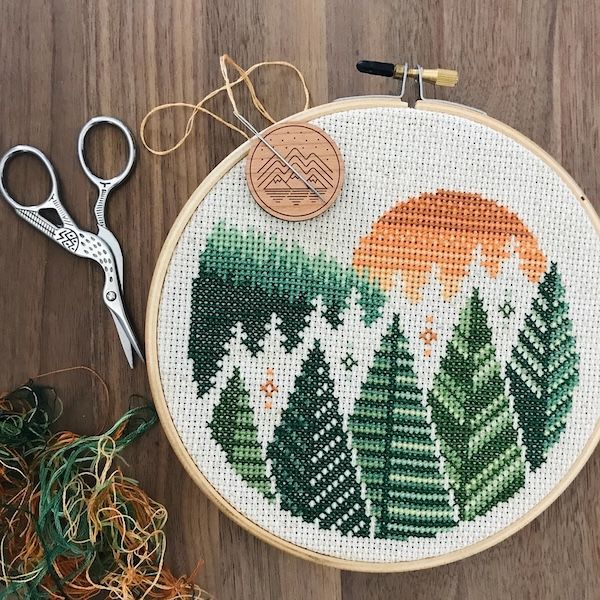 a cross stitch hoop with a forest and sunrise pattern, plus a pair of scissors, thread, and a needle off to the side