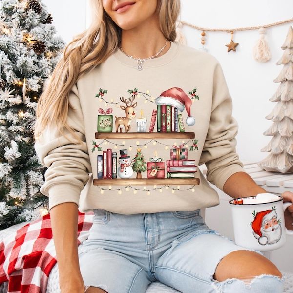 a white woman wearing a tan crewneck sweater with a graphic of two floating bookshelves with books and assorted holiday decorations