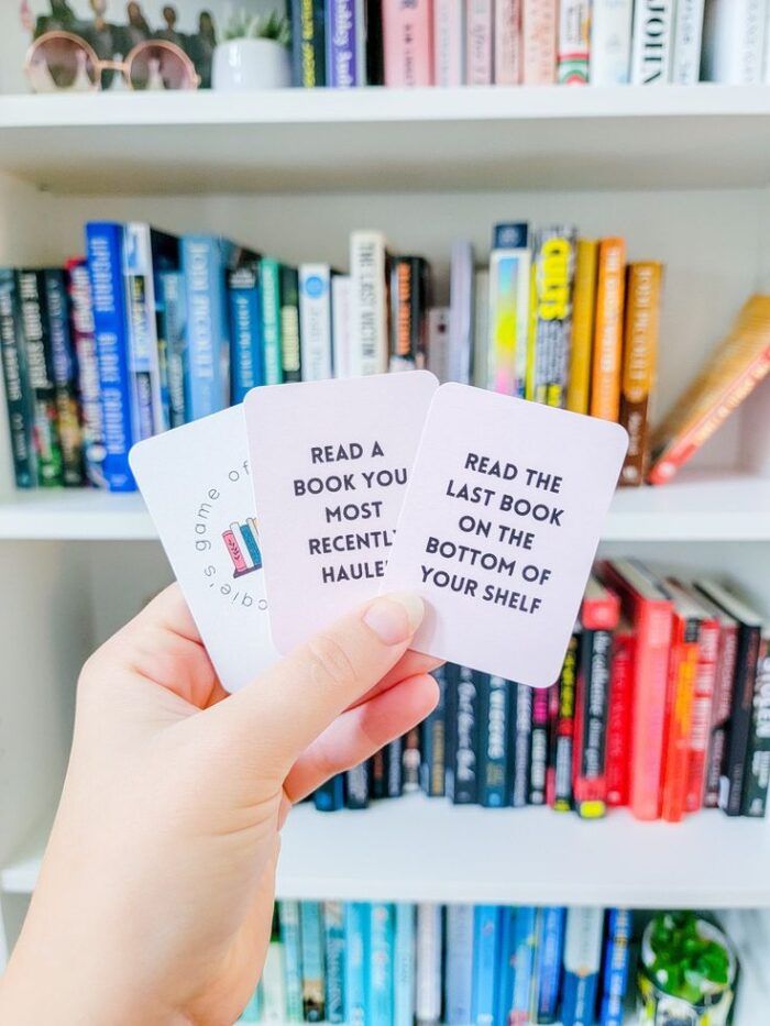 a hand holding cards in front of a rainbow bookcase. cards say things like "read last book on the bottom of your shelf."