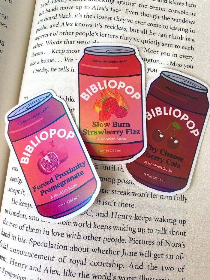 three stickers in the shape of sode cans in reddish hues. The branding matches Olipop sodas, but the cans read Bibilopop with flavors that match book tropes 