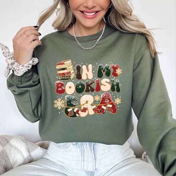 a white woman wearing a light olive green crewneck sweater with text in bubbly letters that reads "in my bookish era" plus graphics of snowflakes and books