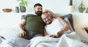 two men reading in bed and smiling, one with his arm around the other