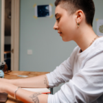 a person with light skin, buzzed hair, and tattoos types on a laptop with small rainbow flags beside them