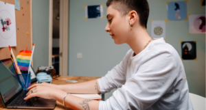 a person with light skin, buzzed hair, and tattoos types on a laptop with small rainbow flags beside them