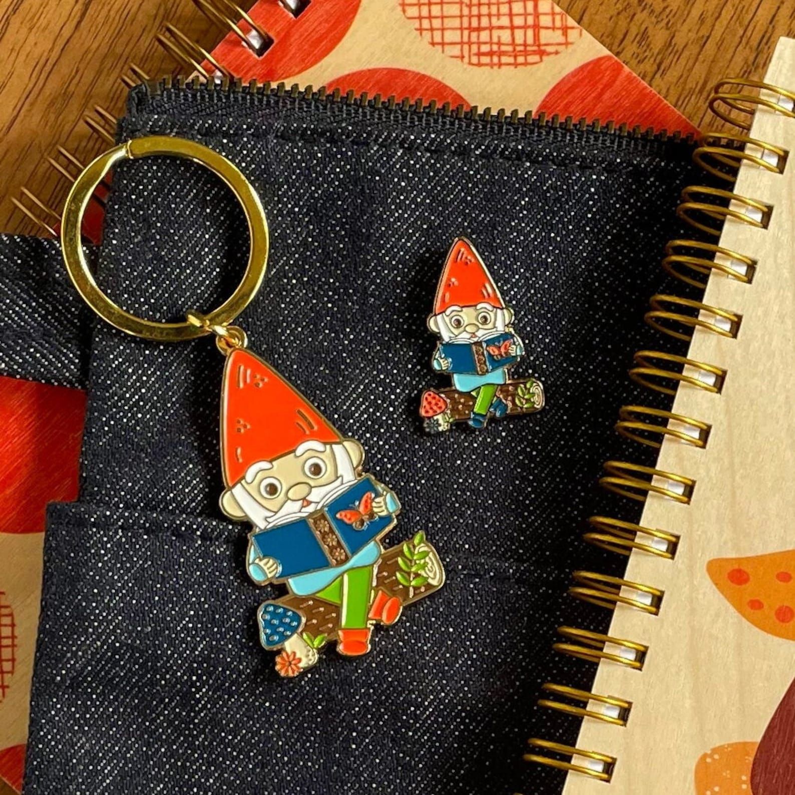 Image of a keychain and a pin on top of a zipper pouch. They are the same image of a colorful nome reading a book. 