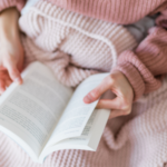 someone wearing a pink sweater reading under a blanket