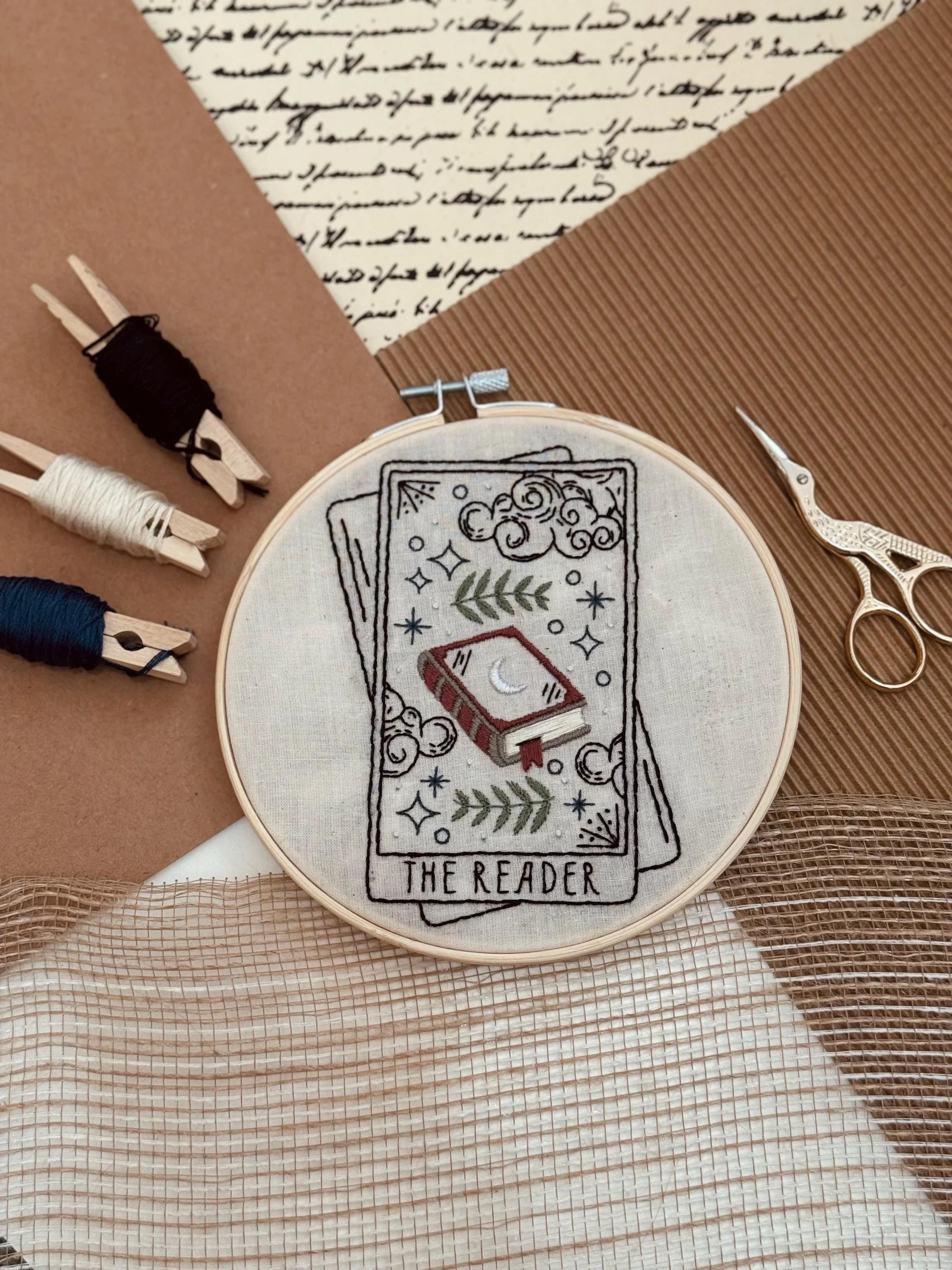 embroidery hoop with an image of a tarot card that says "the reader." It's surrounded by materials in a kit to make the embroidery. 