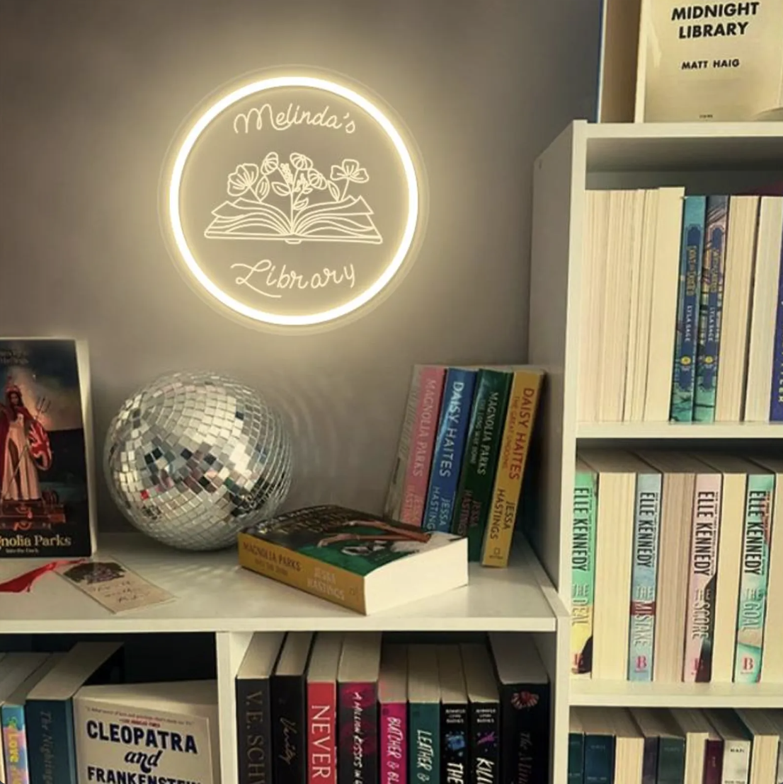 Photo of a circular lit-up sign that says "Melinda's Library" with a floral book design hung on a wall above bookshelves