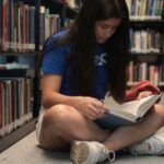 a medium light-skinned young woman sits cross-legged on library floor while reading a book