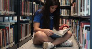 a medium light-skinned young woman sits cross-legged on library floor while reading a book