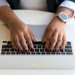 brown-skinned hands with burgundy nails and a gold wrist watch typing on a mac keyboard