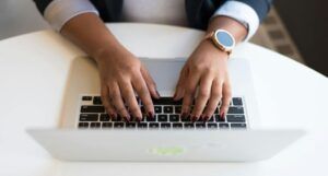 brown-skinned hands with burgundy nails and a gold wrist watch typing on a mac keyboard