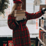 a woman reading a red plaid coat holding a stack of books in a bookstore. A wreath is hanging in the window.