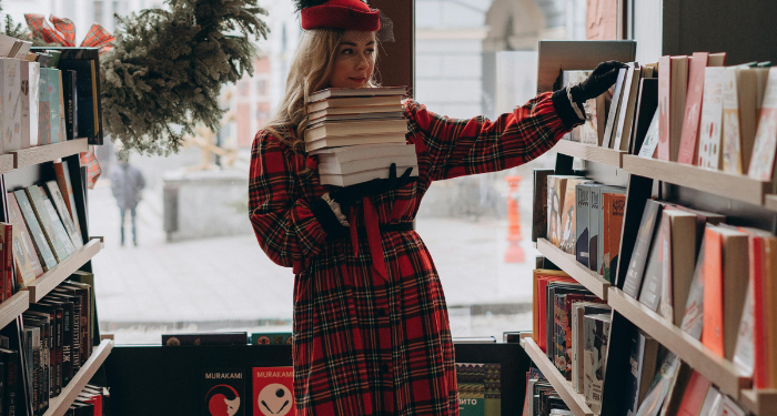 a woman reading a red plaid coat holding a stack of books in a bookstore. A wreath is hanging in the window.